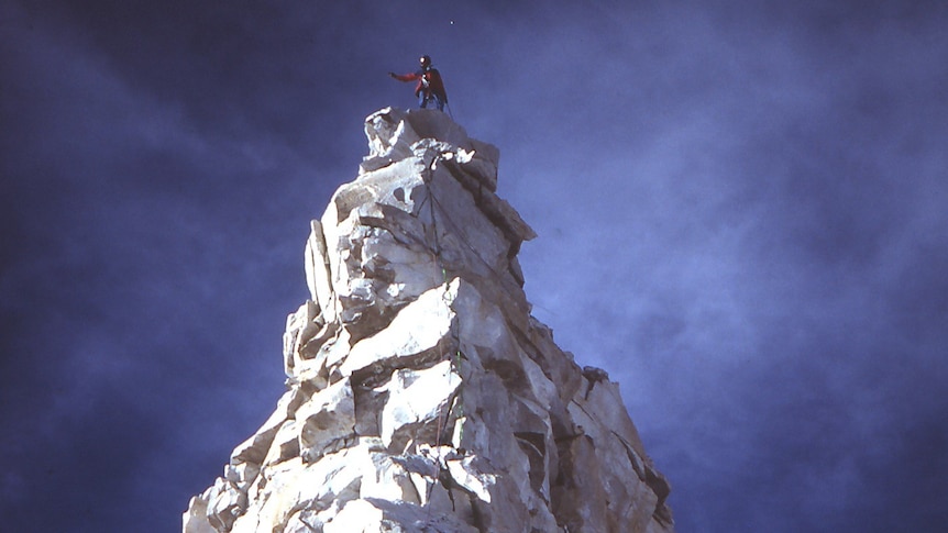 A large, narrow snow-covered mountain with dark sky behind it and a person just visible at its very tip.