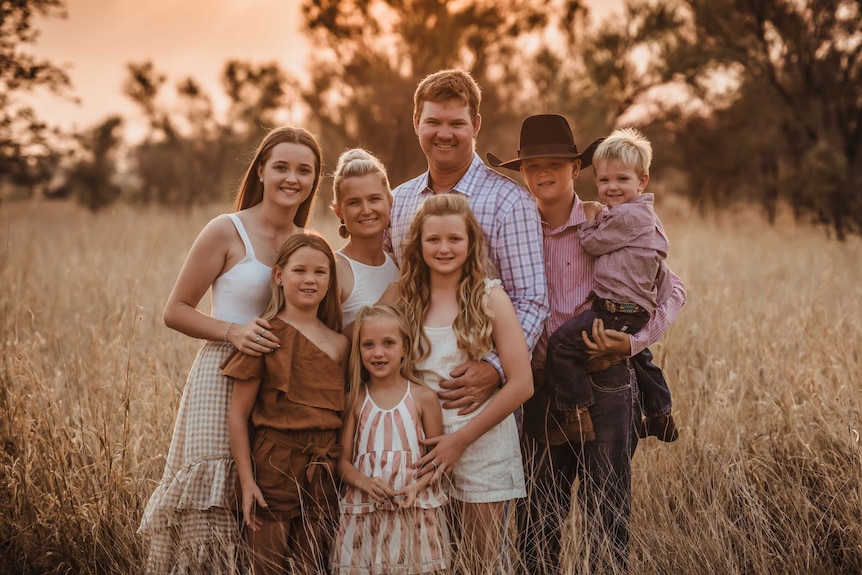 A portrait of the Scott family, parents and six children  at sunset in a wheat paddock, arms around each other in golden light.