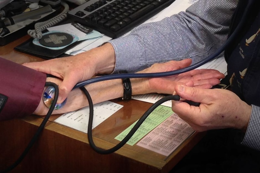 Close up on the arms of a doctor holding a stethoscope to a patient's arms as he takes her blood pressure at a desk.