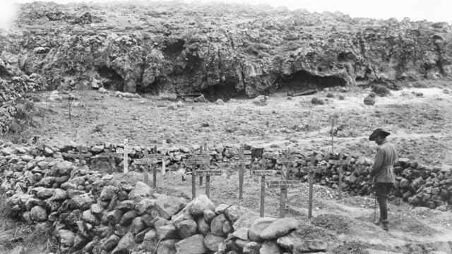A soldier in uniform stands with head bowed before memorial crosses at Gallipoli Peninsula.
