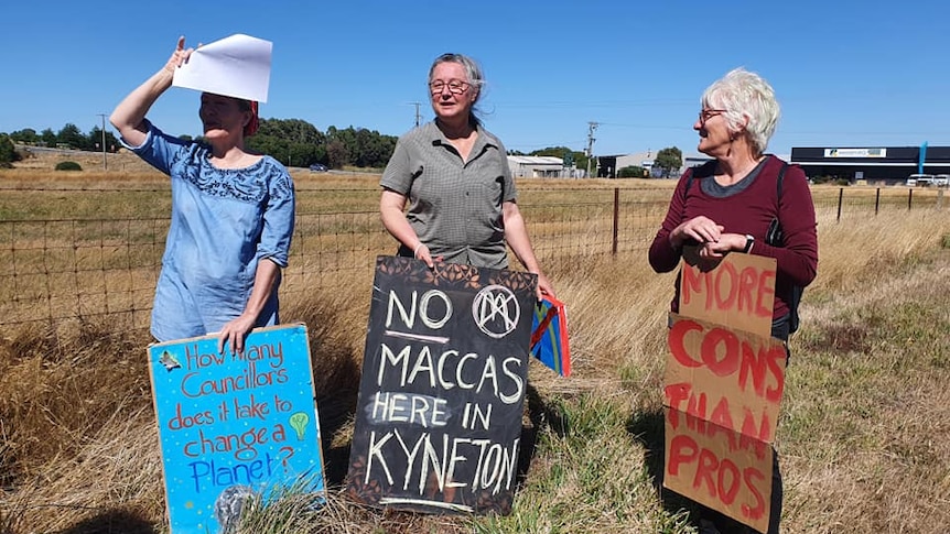 Three residents stand in front of a vacant paddock holding signs saying 'No Maccas here in Kyneton', 'More Cons than pros'. 