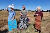 Three residents stand in front of a vacant paddock holding signs saying 'No Maccas here in Kyneton', 'More Cons than pros'. 