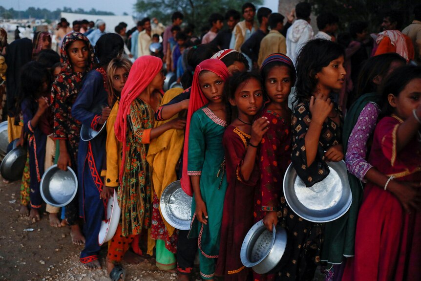 Children with metal bowls queue for food at a camp for flood victims.