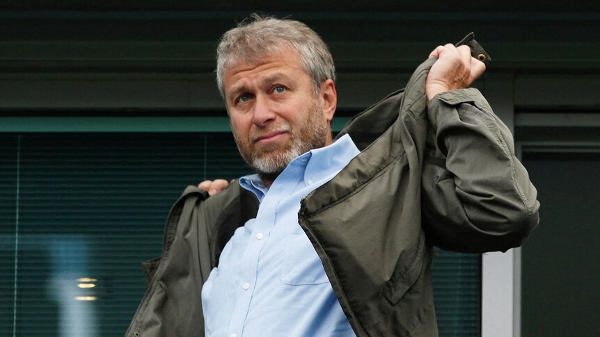 Roman Abramovich, a man with grey hair and stubble, shrugs a khaki jacket on over his blue shirt