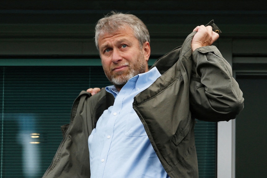 Roman Abramovich, a man with grey hair and stubble, shrugs a khaki jacket on over his blue shirt