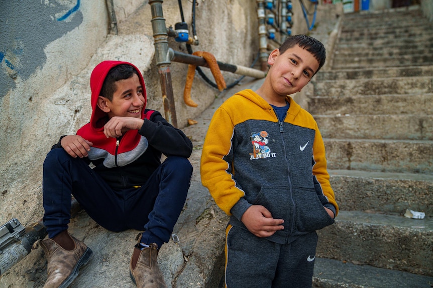 Two young boys, one wearing a red and blue hoodie, the other gold and blue, smile on a set of concrete steps
