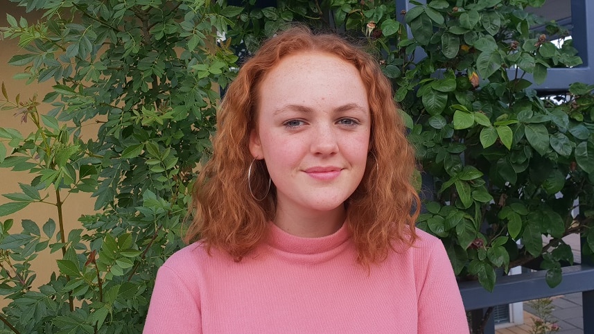 Young woman with red hair and a pink shirt stands in front of greenery