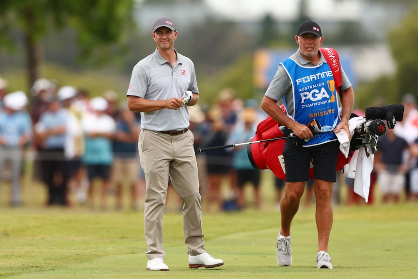 Golfer Adam Scott stands looking down the fairway with his caddie holding his set of clubs during a tournament.