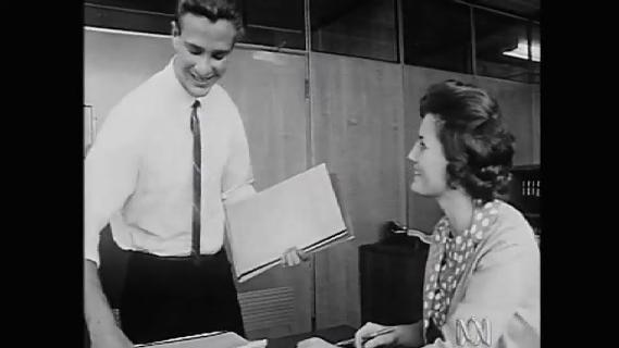 Old photo of man taking papers from office desk, woman sits beside