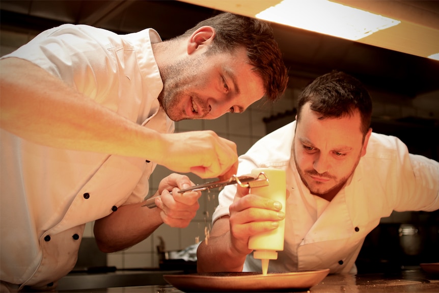 Tom Chiumento grates food while Simon Evans squeezes sauce onto a plate in their kitchen.