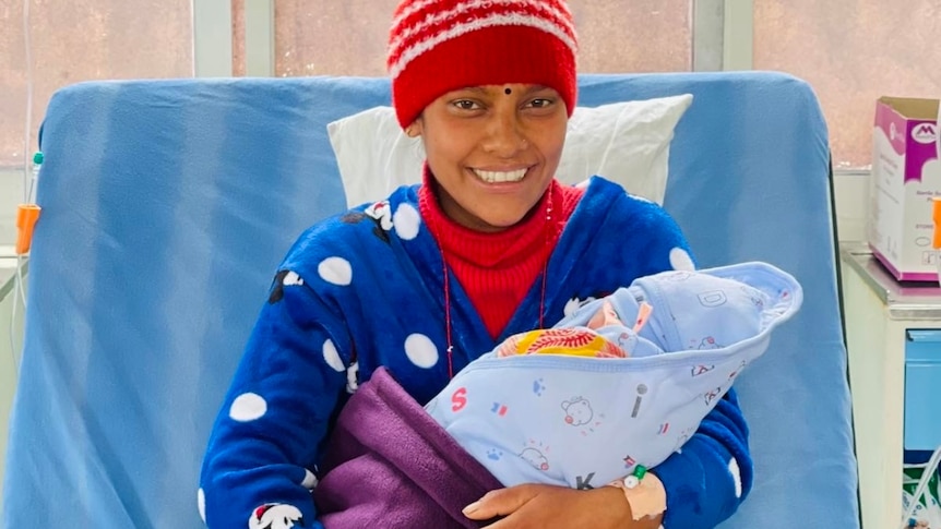 Smiling South Asian woman wears a red and white woollen cap, blue sweater with white polka dots, holds baby, sits hospital bed.