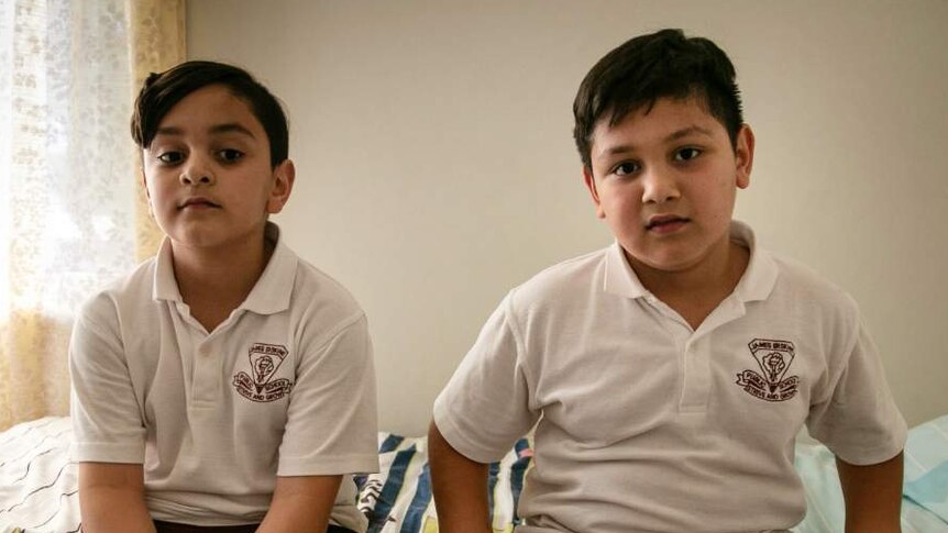 Two young boys sit on a bed, looking at the camera.