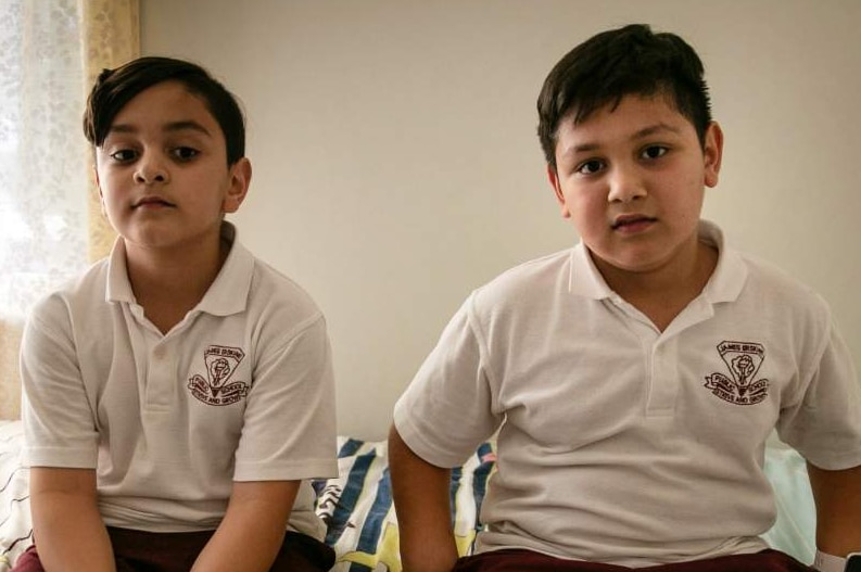 Two young boys sit on a bed, looking at the camera.