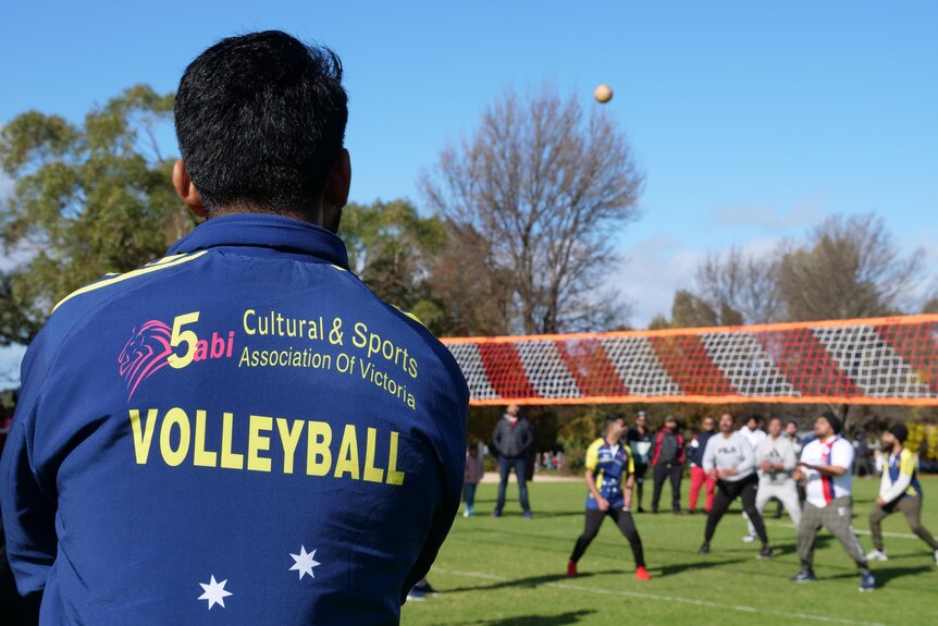 A man wearing a jacket with a logo that reads 'Cultural and Sports Association of Victoria Volleyball' watches volleyball. 