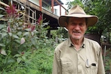 A man wearing a khaki shirt and hat smiles while standing in front of garden.