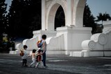 A mother holds the hand of her child who is linked arms with two other kids as they walk near an arch.