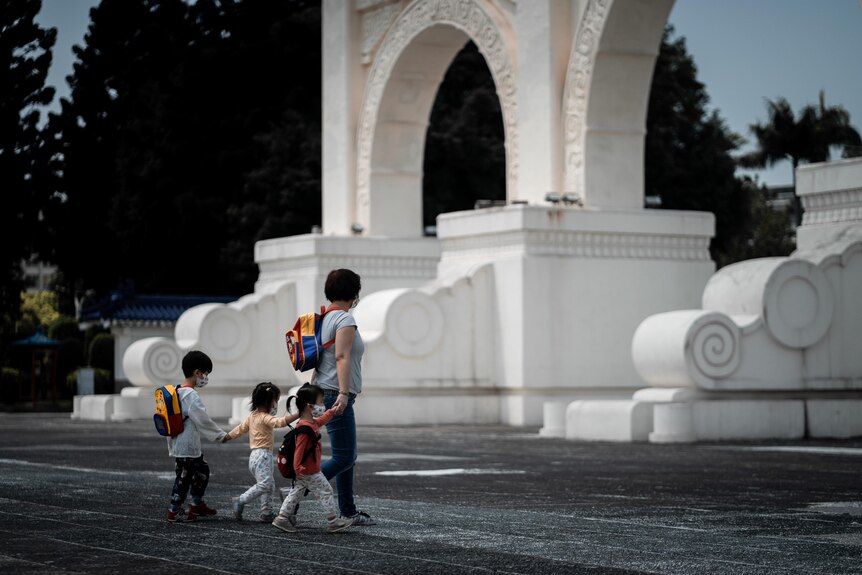 A mother holds the hand of her child who is linked arms with two other kids as they walk near an arch.