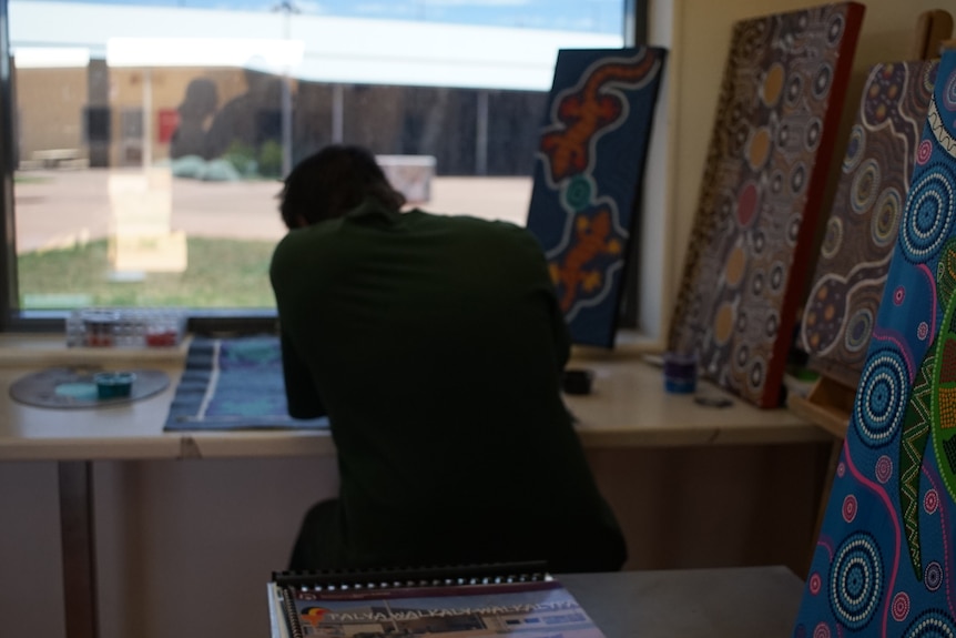 The back of a man painting in front of a window, Indigenous canvas on the side, low buildings with tin roof outside.