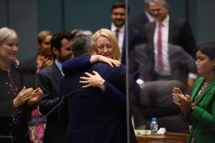 A blonde woman hugging a man in a suit, surrounded by politicians applauding