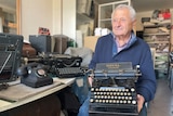 An elderly man sits in a workshop surrounded by old typewriters, with one machine resting on his lap.