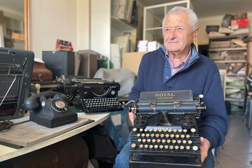 An elderly man sits in a workshop surrounded by old typewriters, with one machine resting on his lap.