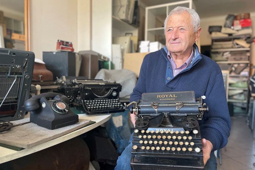 An elderly man sits in a workshop surrounded by old typewriters, with one machine resting on his lap. 