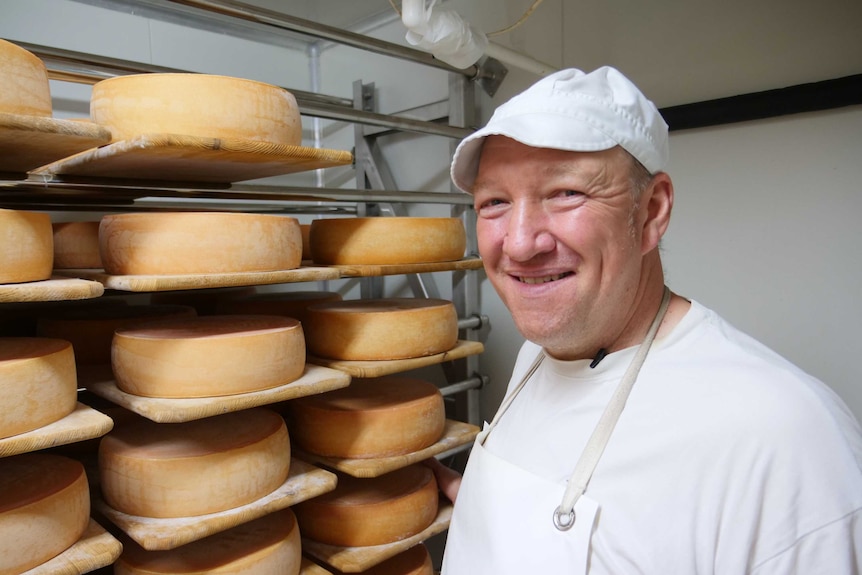 A smiling man in white overalls stands b side several wheels of cheese at a factory