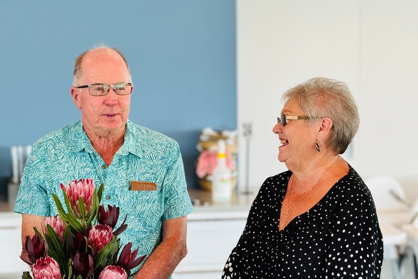 A bunch of protea flowers sit in front of a man in a blue shirt while a woman wearing glasses looks at him laughing. 