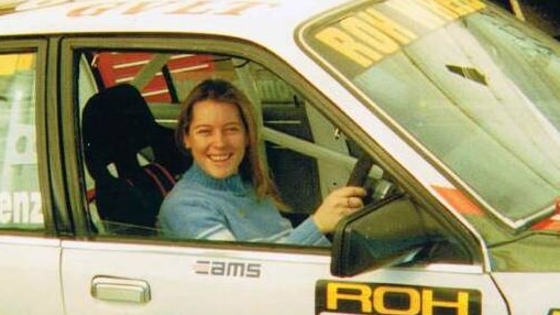 A smiling young woman with long blonde hair sits at the wheel of a racing car.