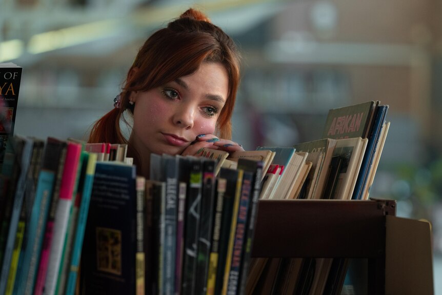 A teenage girl with colour eyeshadow rests her head on a stack of books in the school library.