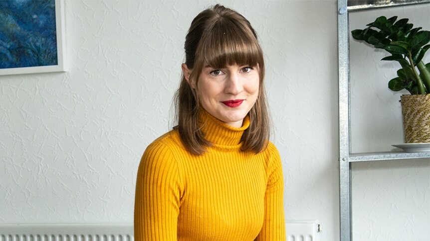 Author Lucia Osborne-Crowley, wearing a yellow turtleneck sweater, sits in front of a bookshelf.
