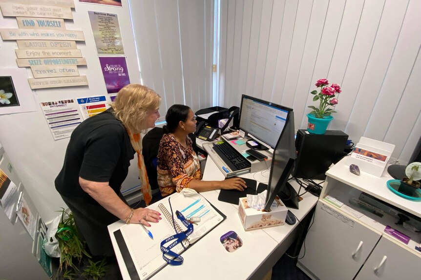 Val Brown stands over the shoulder of a staff member, helping her with work at her desk. 