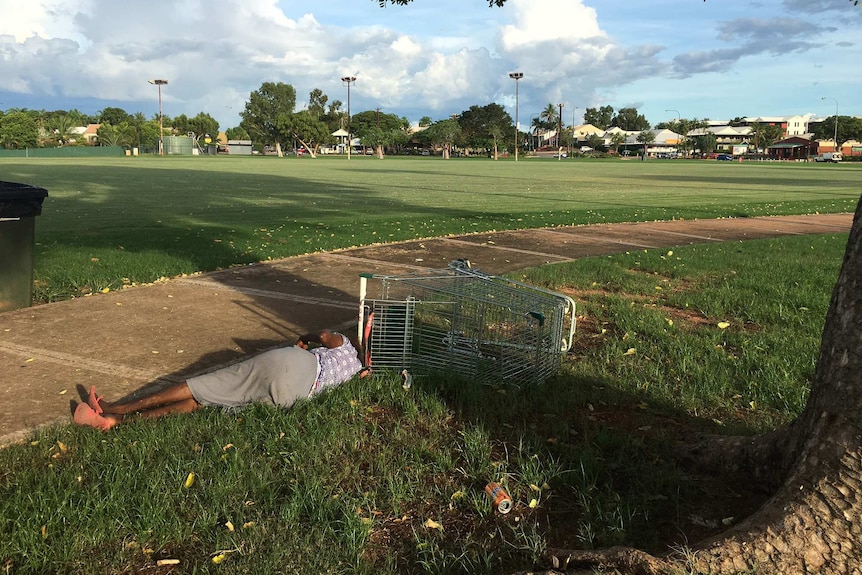 A man lying in the grass next to a footpath and a shopping trolley on its side.
