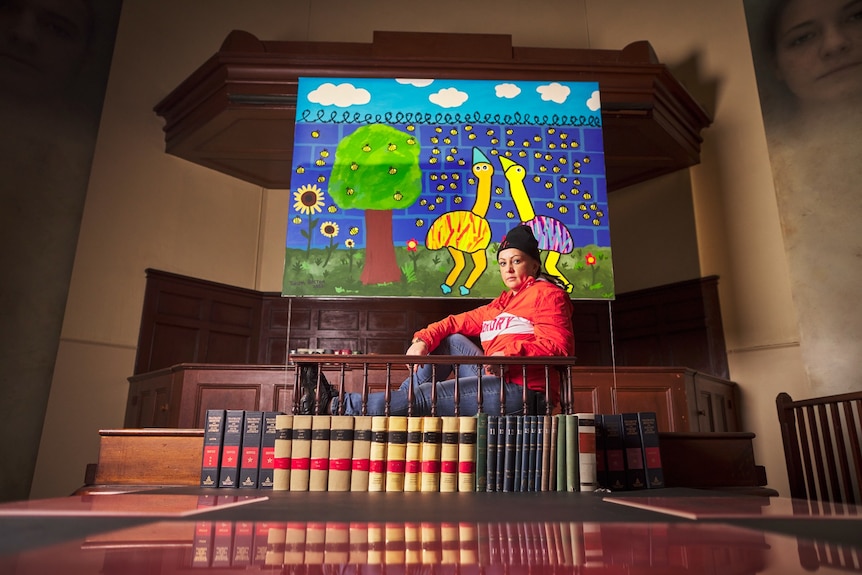 Thelma Coral Beeton sits with her legs stretched out in a court room, screens of colourful art behind her
