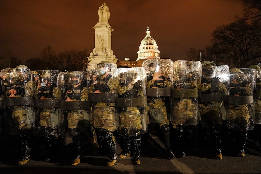 A line of armed people with shields and wearing uniforms, helmets and masks stand outside the Capitol Building.
