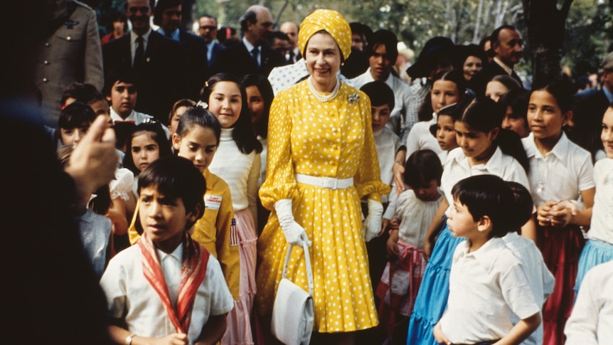 Queen Elizabeth, wearing a yellow with white polka dots dress and matching turban, stands with a group of children