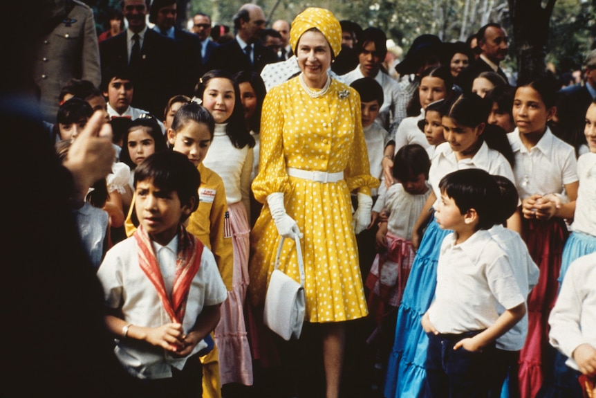 Queen Elizabeth, wearing a yellow with white polka dots dress and matching turban, stands with a group of children