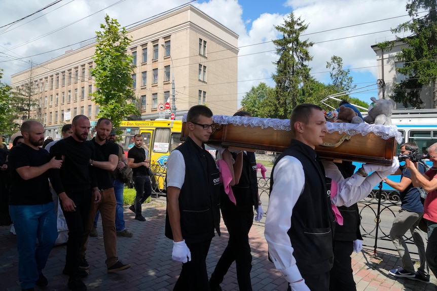 Four men carry a small wooden coffin in the street. Three men in black crying walk behind. 