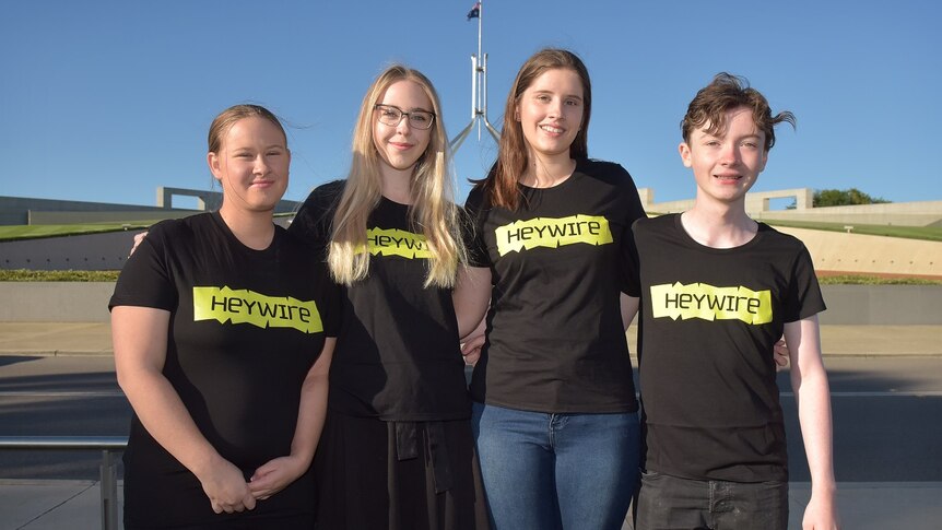 Four young people in Heywire shirts stand in front of Parliament House with arms around each other.