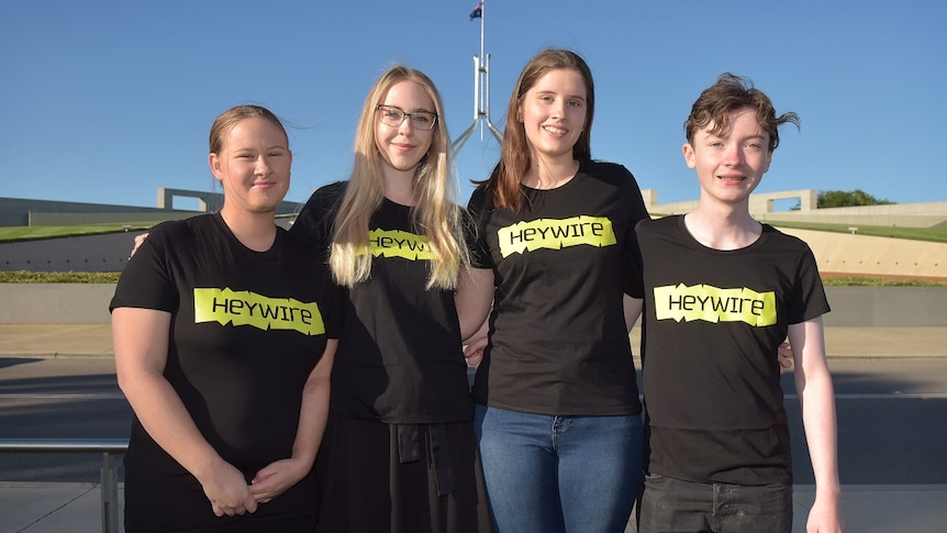 Four young people in Heywire shirts stand in front of Parliament House with arms around each other.