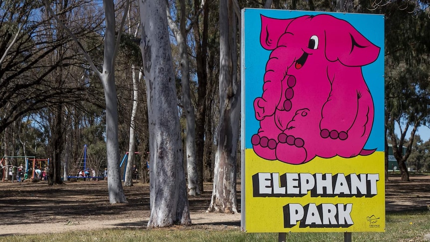 A sign with a big pink elephant on it saying Elephant Park with trees and a playground in the background