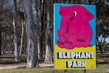 A sign with a big pink elephant on it saying Elephant Park with trees and a playground in the background
