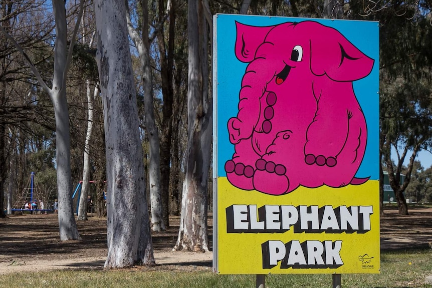 A sign with a big pink elephant on it saying Elephant Park with trees and a playground in the background