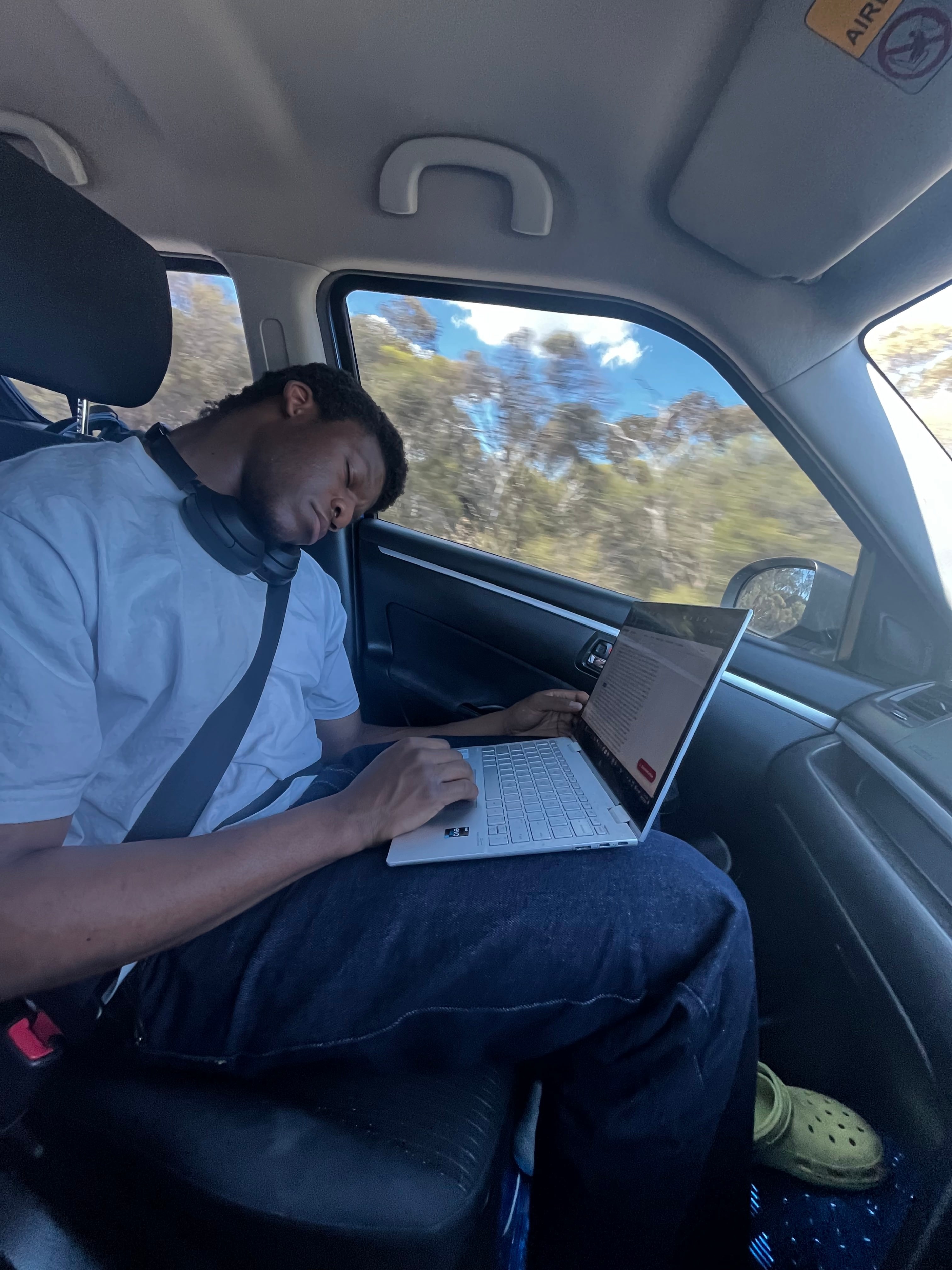 Um homem de camiseta dormindo no banco do passageiro de um carro com seu laptop no colo.