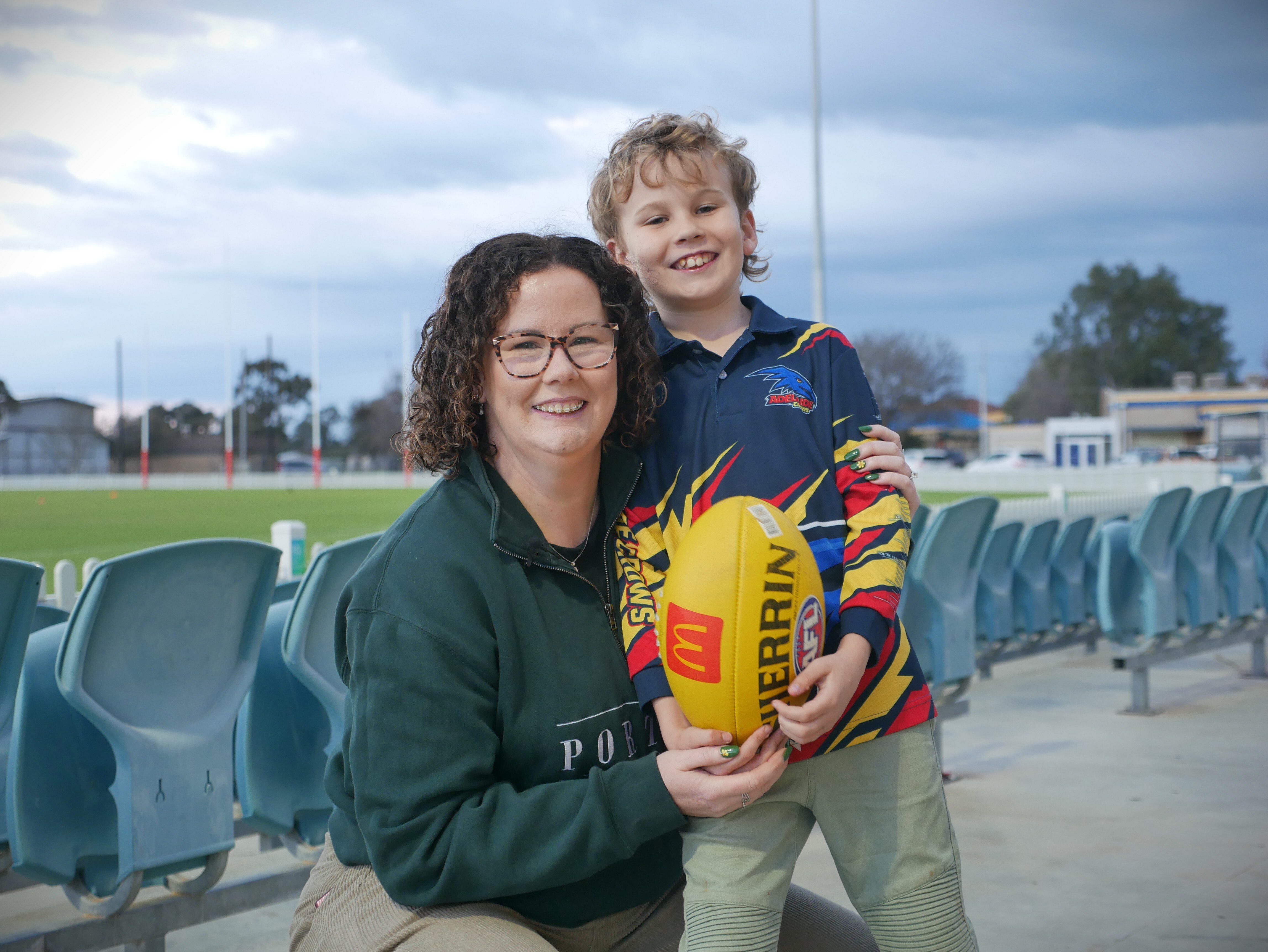 A mother smiling and hugging a young boy in Crows top holding a football next to seats at an oval
