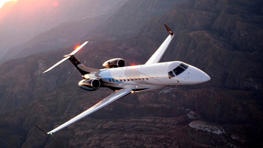 You view a white private jet flying over mountainous terrain at sunset.