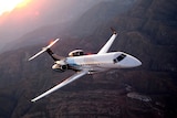 You view a white private jet flying over mountainous terrain at sunset.