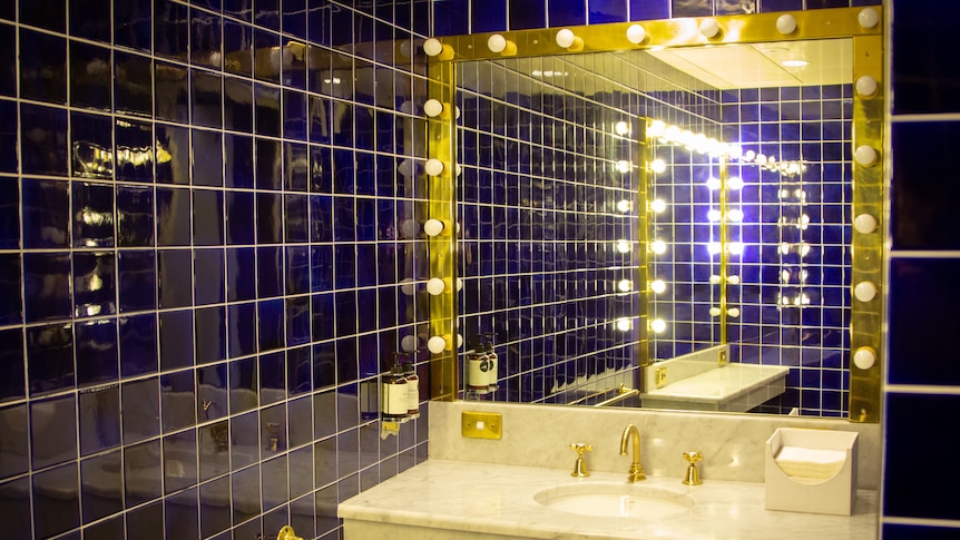 A bathroom with royal blue tiles and a vanity with dressing room lights that leave a gold hue