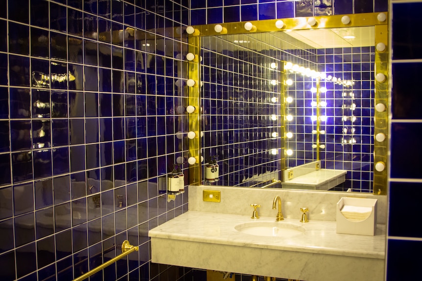 A bathroom with royal blue tiles and a vanity with dressing room lights that leave a gold hue