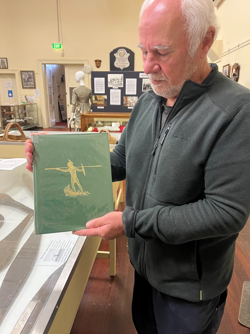 An older man holds a book with an embossed image of a First Nations warrior on the front cover